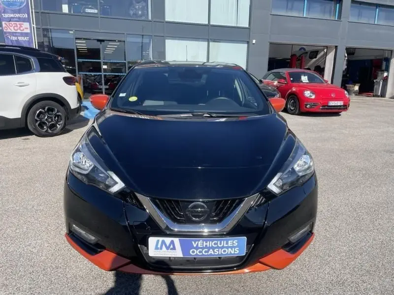 Vue avant d'une Nissan Micra noire avec détails orange, badge Nissan visible, stationnée devant un bâtiment commercial.