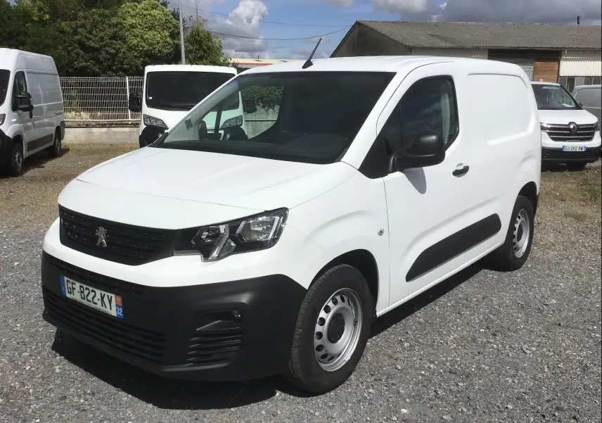 Vue 3/4 avant d’un utilitaire Peugeot Partner blanc avec pare-chocs noirs et jantes acier, stationné sur un sol gravillonné.