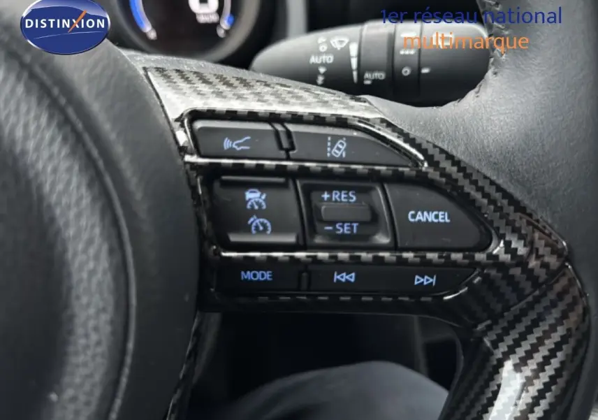 Gros plan sur les commandes au volant en carbone du Toyota Yaris Cross 116h Design noir, avec régulateur de vitesse et mode audio.