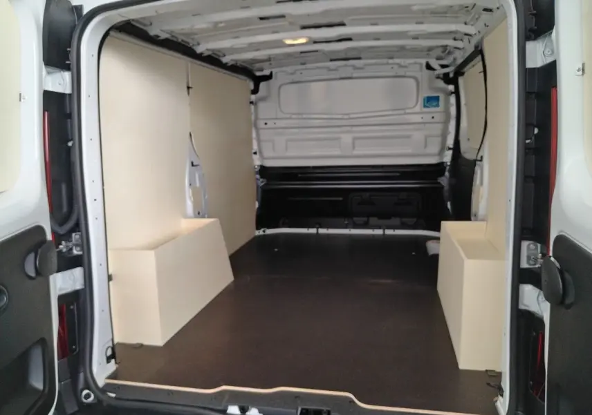 Vue intérieure arrière du fourgon blanc Nissan Primastar 2025 avec aménagement bois et plancher noir.