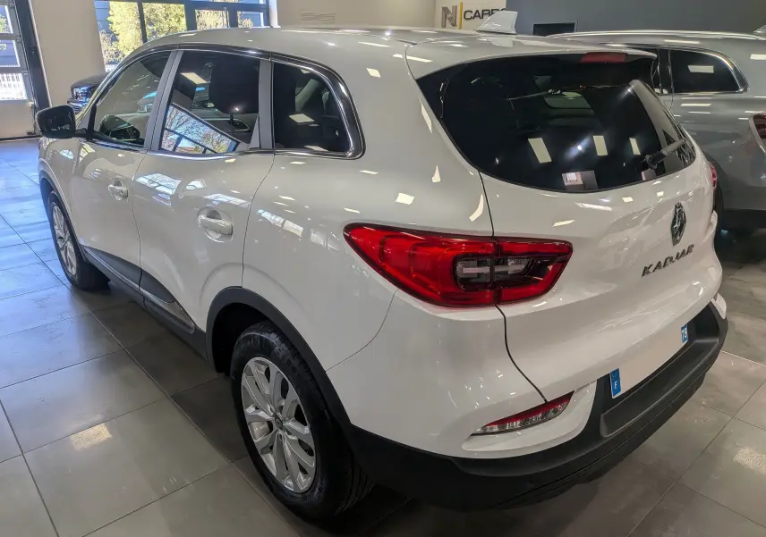Vue 3/4 arrière droite d'un Renault Kadjar blanc Glacier, avec feux arrière rouges et vitres teintées, en intérieur showroom.