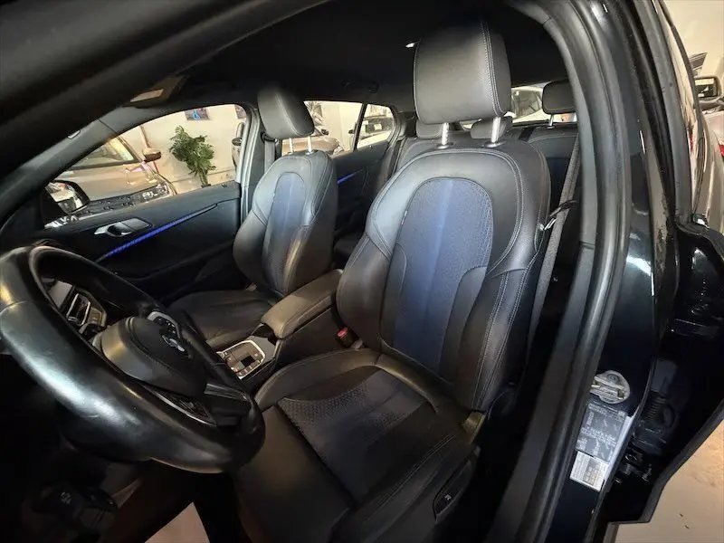 Vue intérieure côté conducteur d'une BMW Série 1 noire, sièges en cuir noir avec surpiqûres bleues et éclairage d'ambiance bleu.