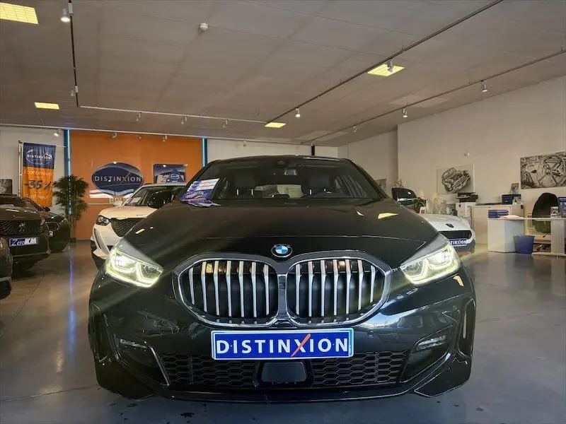 Vue avant d'une BMW Série 1 noire métal avec calandre caractéristique et phares allumés en showroom.