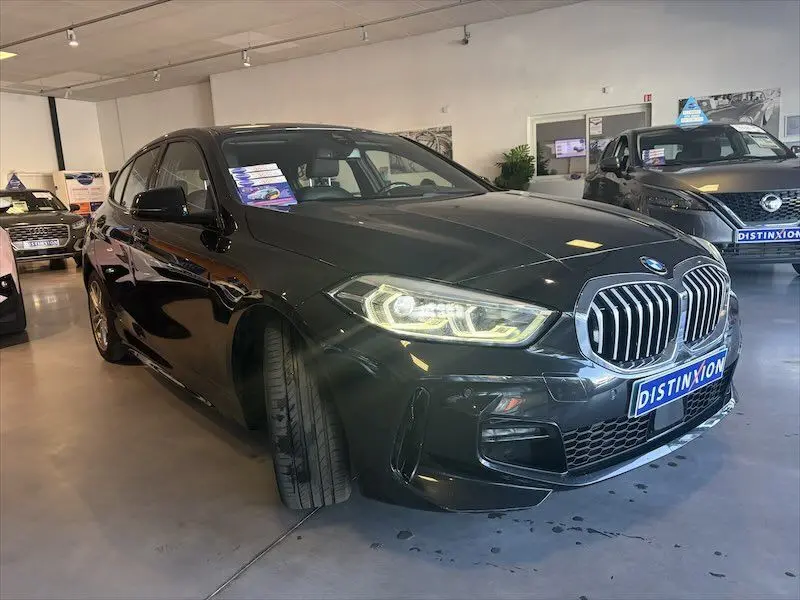 BMW Série 1 noire métal vue 3/4 avant gauche, avec calandre imposante et phares LED allumés en showroom.
