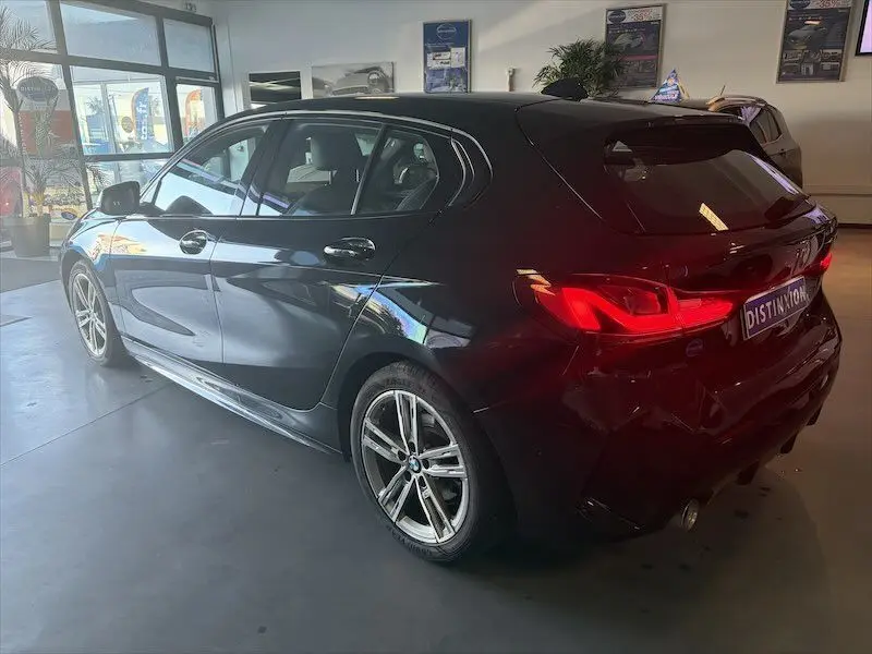 BMW Série 1 noire métal vue 3/4 arrière côté gauche, avec feux arrière allumés en intérieur showroom.