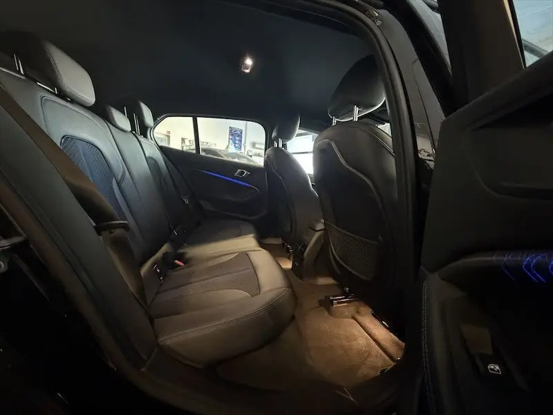 Vue intérieure arrière côté gauche d'une BMW Série 1 noire métal avec sièges en cuir noir et éclairage d'ambiance bleu.