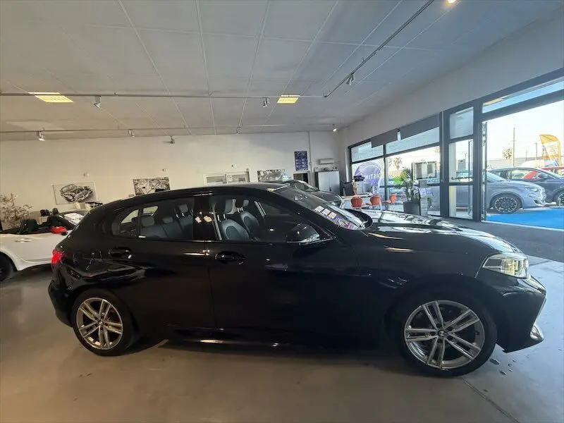 BMW Série 1 noire métal vue de profil côté gauche dans un showroom lumineux avec intérieur visible.