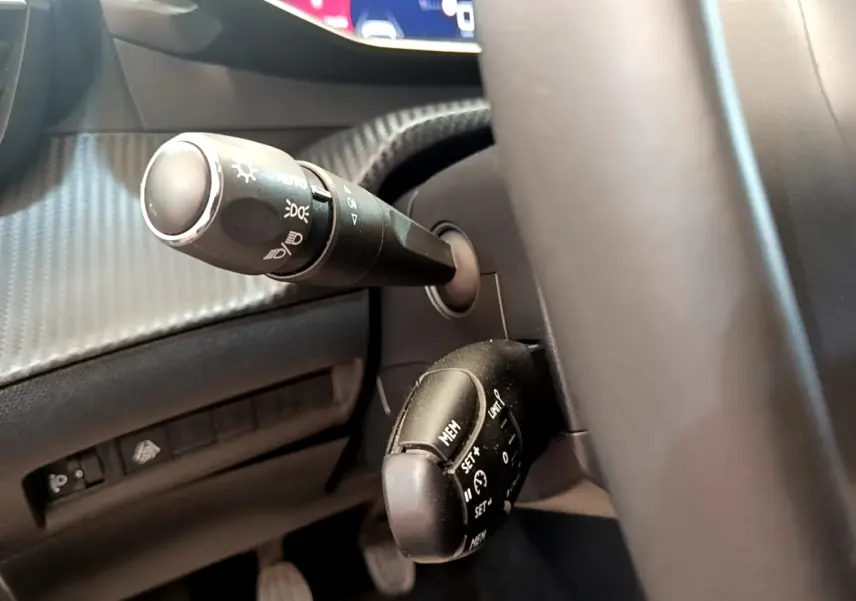 Gros plan sur la colonne de direction à gauche du volant cuir d’une Peugeot 208 blanche, avec commandes de phares et régulateur.