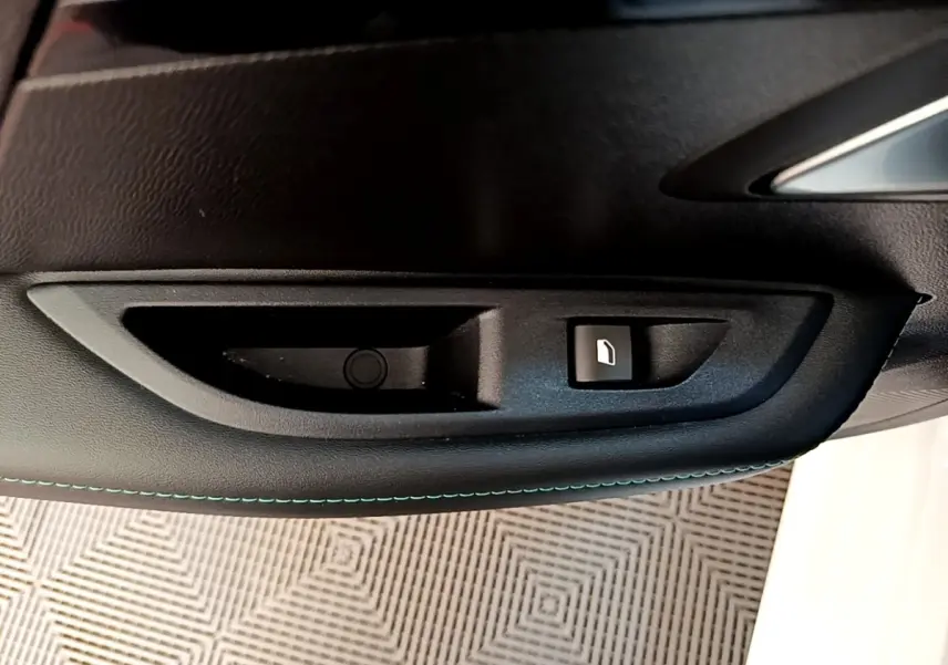 Gros plan sur la commande de lève-vitre côté conducteur d'une Peugeot 208 blanche, avec surpiqûres bleues sur la porte.