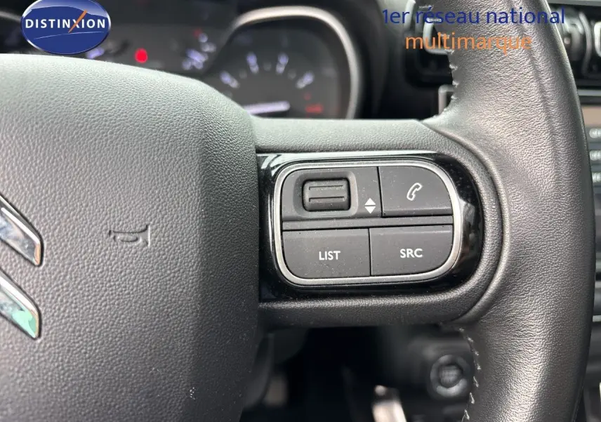 Gros plan sur les commandes à droite du volant en cuir noir du Citroën C3 Aircross Shine Gris Platinium, tableau de bord flou en arrière-plan.