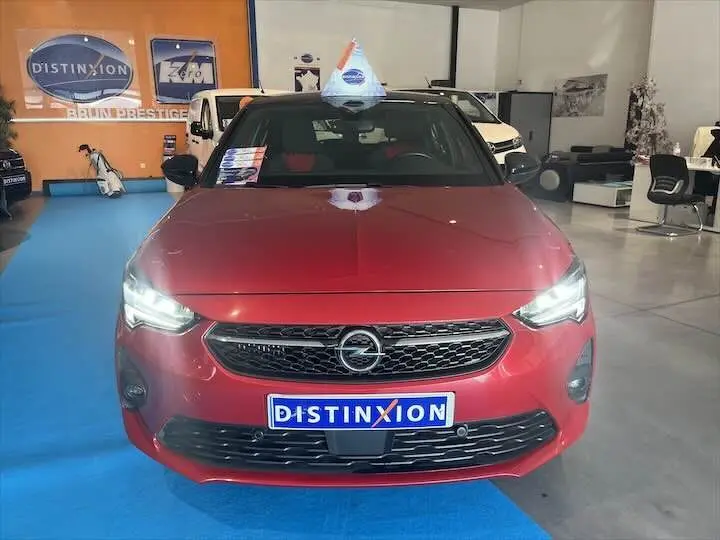 Vue frontale d'une Opel Corsa rouge Kardio avec phares allumés et plaque Distinxion en concession.