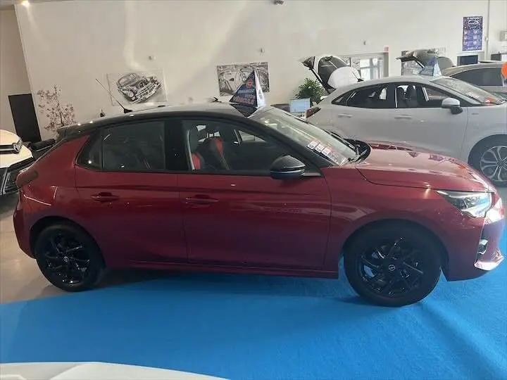 Profil côté gauche d'une Opel Corsa rouge Kardio 2021 avec jantes noires, exposée en intérieur showroom.