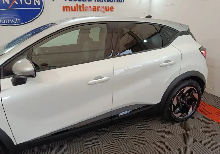 Profil côté gauche du Renault Captur blanc 2025 avec jantes noires et vitres teintées en intérieur showroom.