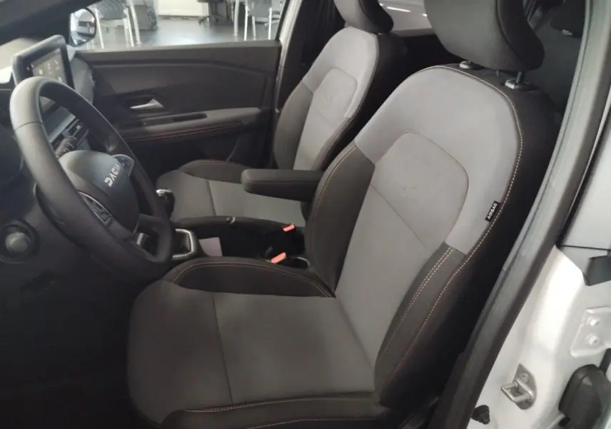 Vue intérieure côté conducteur du Dacia Jogger 2025 blanc, sièges gris avec surpiqûres marron et volant multifonction.