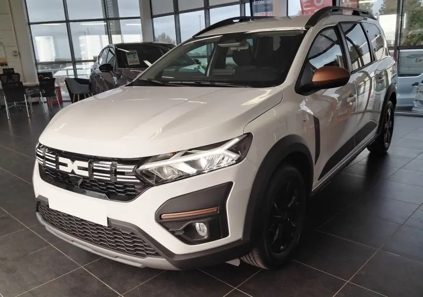Vue 3/4 avant d'un Dacia Jogger blanc 2025 avec barres de toit modulables et rétroviseurs cuivrés en showroom.