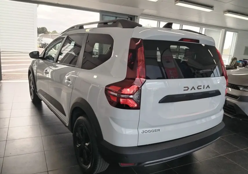Vue 3/4 arrière d'un Dacia Jogger blanc avec barres de toit modulables et feux arrière LED en intérieur showroom.