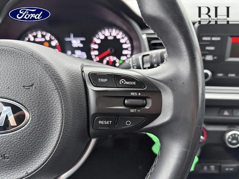 Gros plan sur le volant cuir noir de la KIA Rio 2018 avec commandes de régulateur de vitesse et tableau de bord flou en arrière-plan.