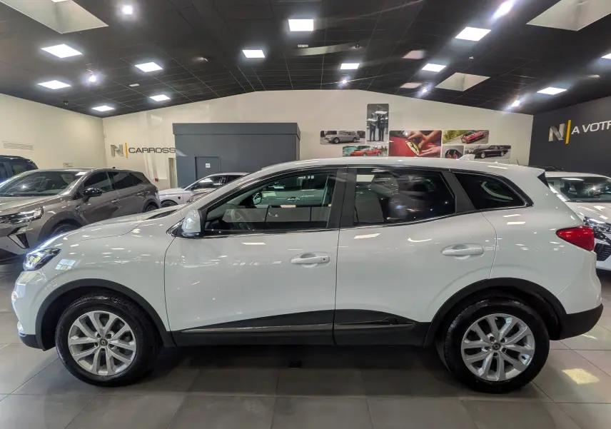 Profil côté gauche d'un Renault Kadjar Blanc Glacier 2021 en showroom, avec jantes alliage et détails noirs sur les bas de caisse.