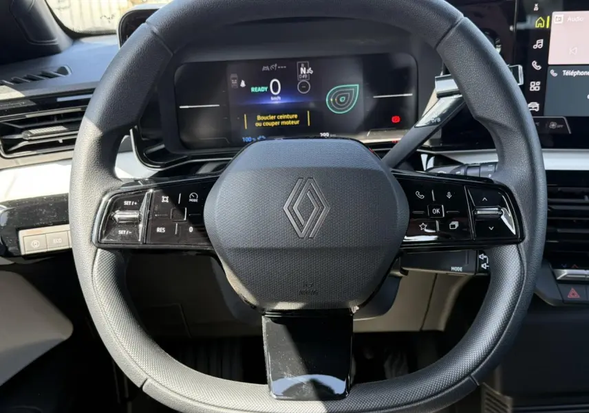 Vue rapprochée du volant noir grainé avec logo Renault, tableau de bord numérique et commandes multifonctions visibles.