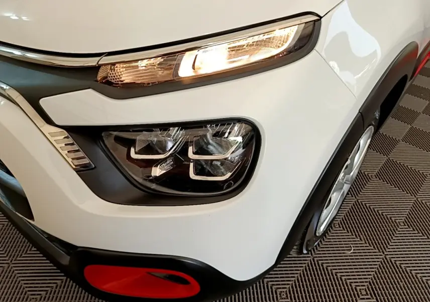 Gros plan sur le phare avant droit allumé d'une Citroën C3 blanche, avec détail rouge sur le pare-chocs noir.