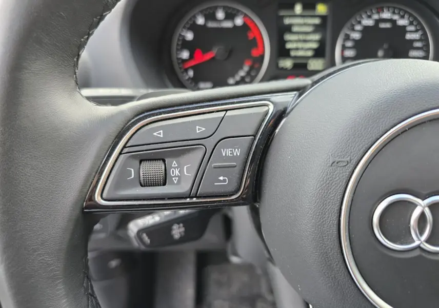 Gros plan sur les commandes à gauche du volant cuir d'un Audi Q2 noir mythic, avec compteurs en arrière-plan.