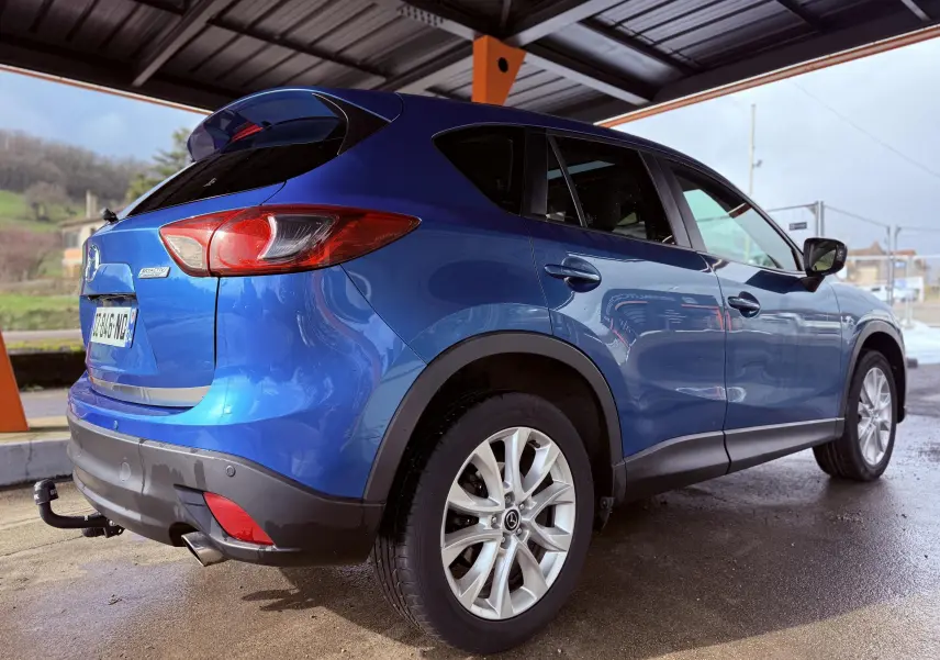 Vue 3/4 arrière droite d'un Mazda CX-5 bleu 2013 avec attelage et jantes argentées sous abri.