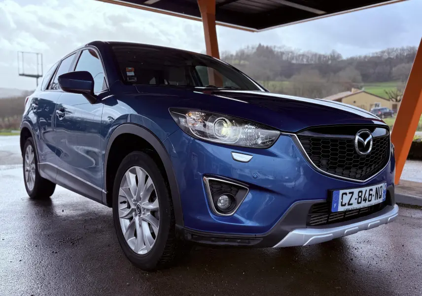 Vue 3/4 avant gauche d'un Mazda CX-5 bleu 2013 avec calandre noire et feux allumés sous abri.