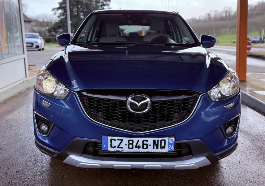 Vue de face du Mazda CX-5 bleu 2013 avec calandre noire et phares allumés sous un abri.