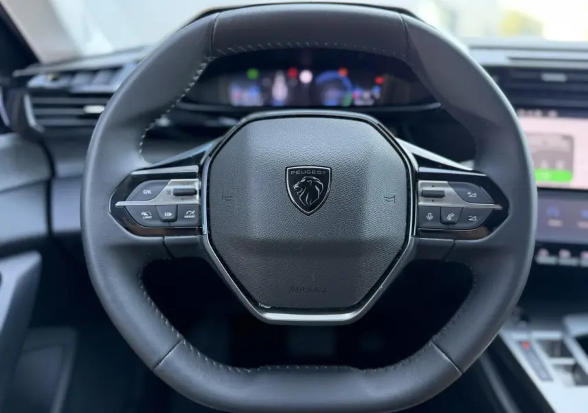 Gros plan sur le volant cuir noir de la Peugeot 308 Hybrid 2025 avec commandes intégrées et logo central lion.