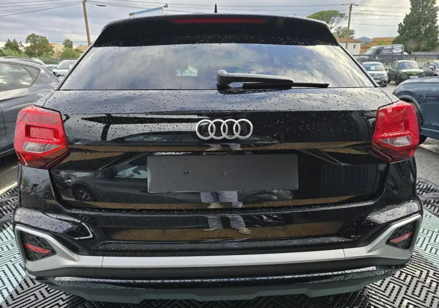 Vue arrière d'un Audi Q2 noir mythic avec feux rouges et lunette arrière surteintée sous la pluie.