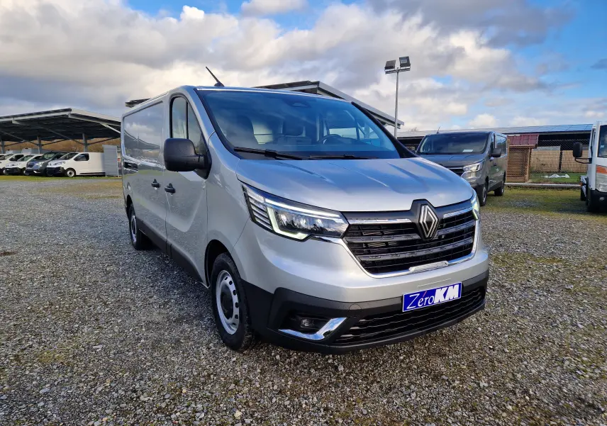 Vue 3/4 avant droit d'un Renault Trafic Fourgon gris Highland avec calandre noire et feux LED allumés sur un parking.