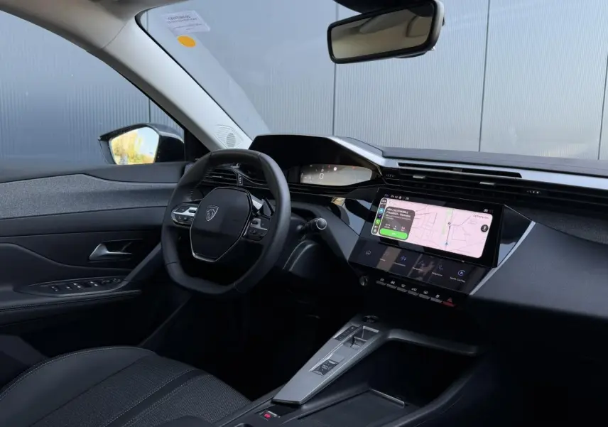 Intérieur Peugeot 308 Hybrid 2025 vu côté conducteur, tableau de bord noir avec écran tactile 10 pouces et volant compact.