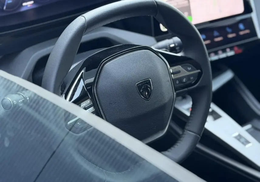 Gros plan sur le volant cuir noir de la Peugeot 308 Hybrid 145, avec tableau de bord numérique et écran tactile visible.