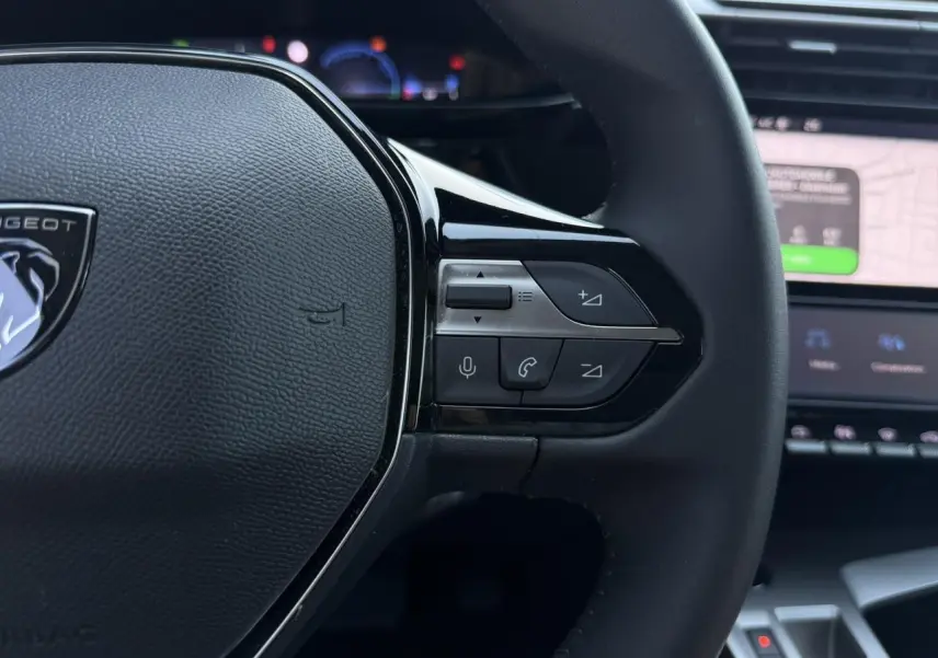 Gros plan sur le volant noir de la Peugeot 308 Hybrid 2025 avec commandes intégrées et écran tactile en arrière-plan.