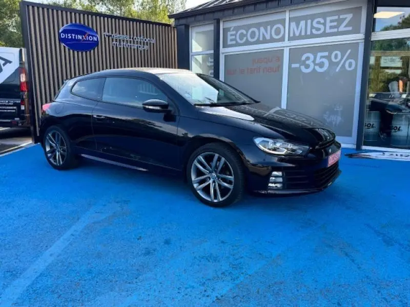 Volkswagen Scirocco noir intense en 3/4 avant droit, jantes alu et calandre distinctive sur parking bleu.