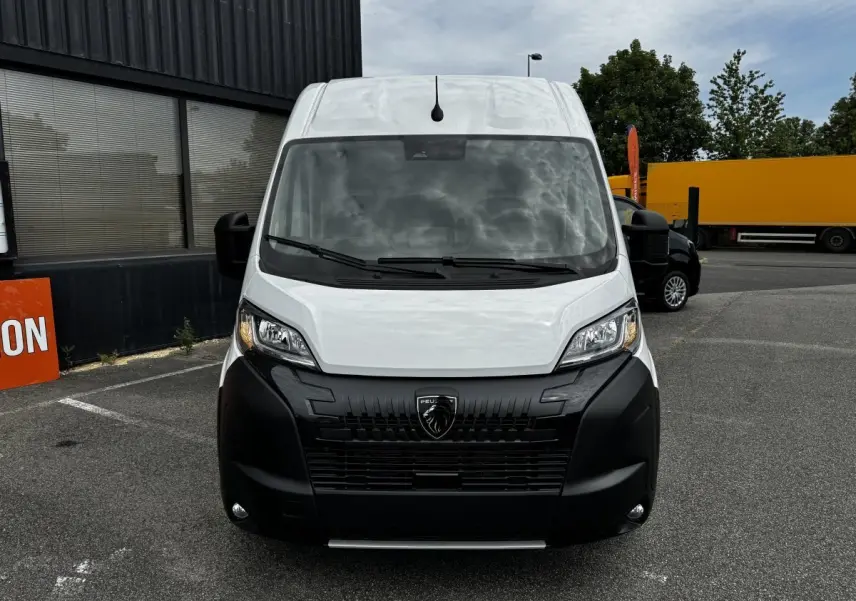 Vue avant d’un Peugeot Boxer Fourgon blanc avec calandre noire et phares allumés sur un parking extérieur.