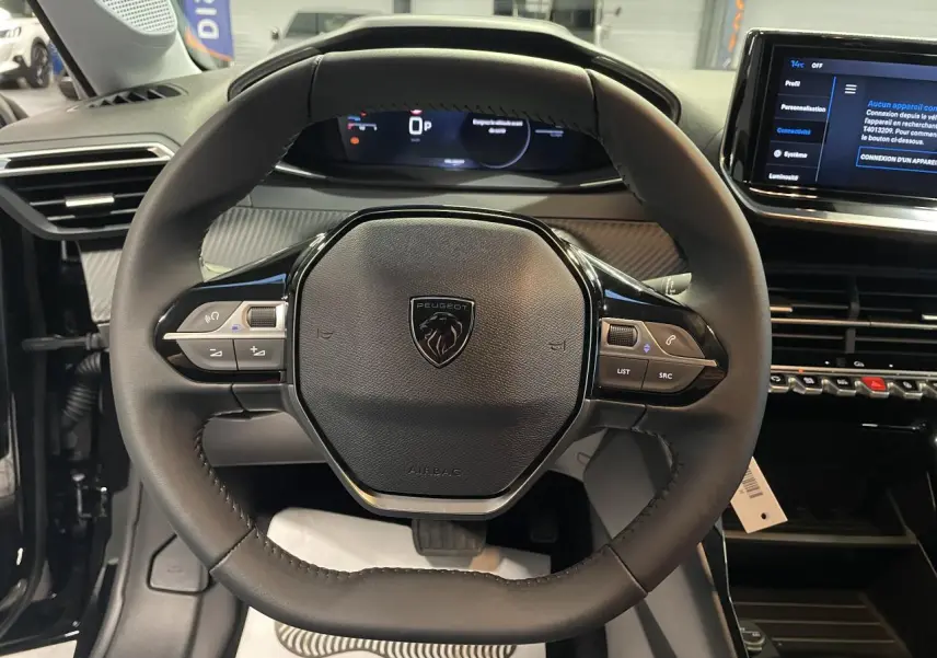Vue rapprochée du volant cuir noir de la Peugeot 208 1.2i Hybrid avec tableau de bord numérique et commandes intégrées.