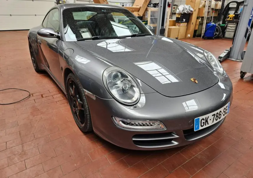 Porsche 911 Carrera 4S gris météore vue 3/4 avant gauche dans un atelier avec jantes noires et phares ronds.