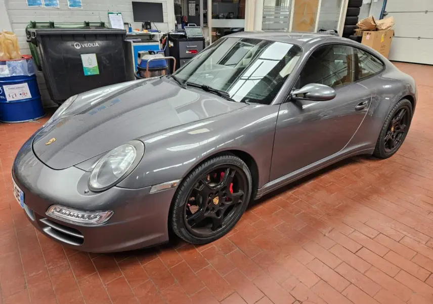 Porsche 911 Carrera 4S gris météore vue 3/4 avant droit, jantes noires Carrera Sport 19 pouces et freins rouges visibles.