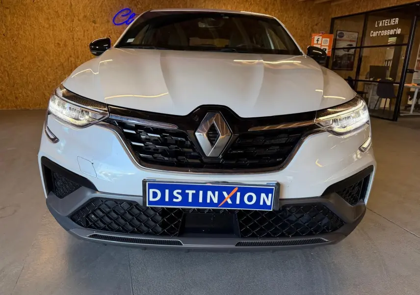 Vue avant d'un Renault Arkana blanc nacré avec calandre noire et phares LED allumés en intérieur showroom.