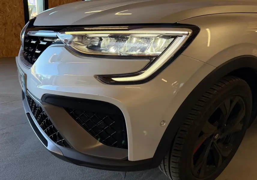 Gros plan sur l'avant droit blanc nacré du Renault Arkana 1.6 E-Tech hybrid 145 RS LINE avec phares LED et jante alu noire.
