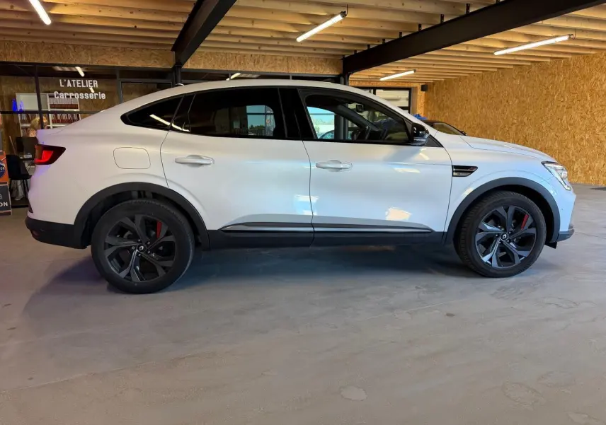 Vue de profil côté gauche d'un Renault Arkana blanc nacré avec jantes alu noires et étriers rouges, stationné en intérieur.