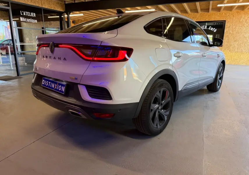 Vue 3/4 arrière droite d'une Renault Arkana blanc nacré avec feux arrière allumés et jantes alu noires.