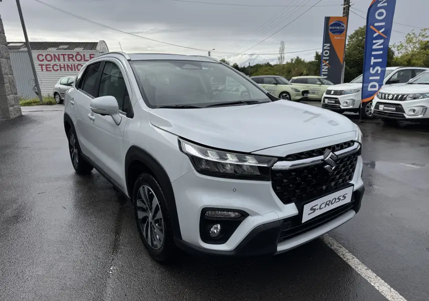 Suzuki S-Cross blanc vue 3/4 avant droit, avec calandre noire et phares LED, sur parking humide devant un garage.