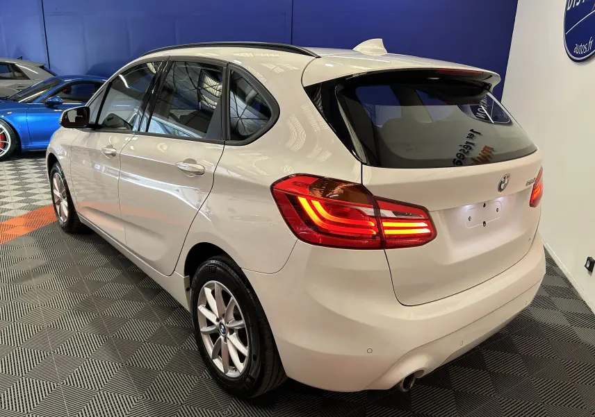 BMW Série 2 Active Tourer blanche vue en 3/4 arrière droit, feux arrière allumés dans un showroom intérieur.