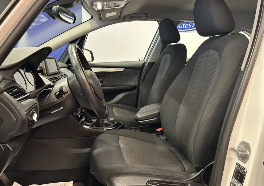 Vue intérieure côté conducteur de la BMW Série 2 Active Tourer blanche, montrant les sièges en tissu noir avec appuie-têtes et volant multifonction.