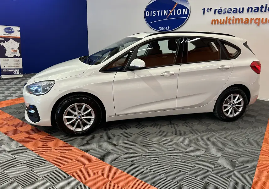 BMW Série 2 Active Tourer 216d blanc vue de profil côté gauche en intérieur, avec jantes alliage et feux allumés.
