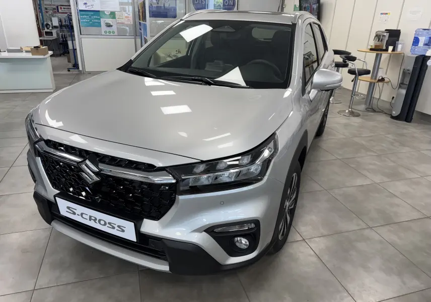 Vue 3/4 avant d'un Suzuki S-Cross gris 2025 en showroom, avec calandre noire et phares LED distinctifs.