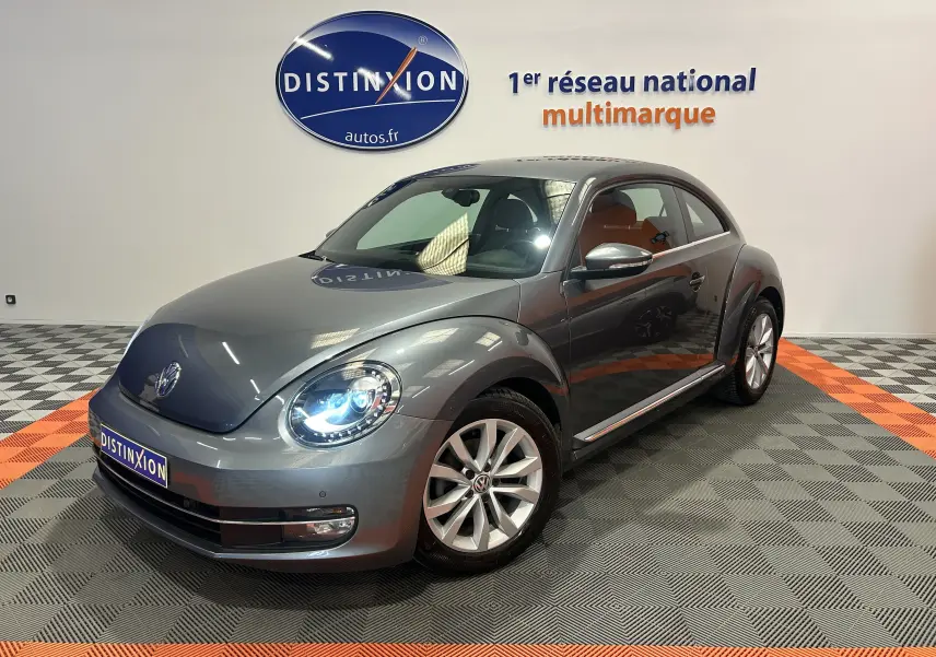 Volkswagen Coccinelle gris vue 3/4 avant droit avec phares allumés dans showroom lumineux.