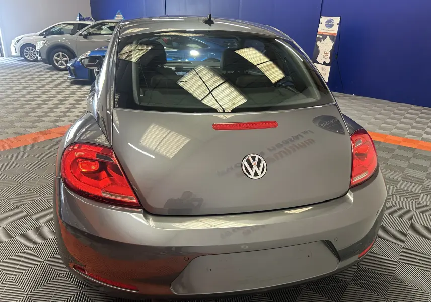 Vue arrière d'une Volkswagen Coccinelle gris métallisé 2016 dans un showroom avec feux arrière rouges bien visibles.